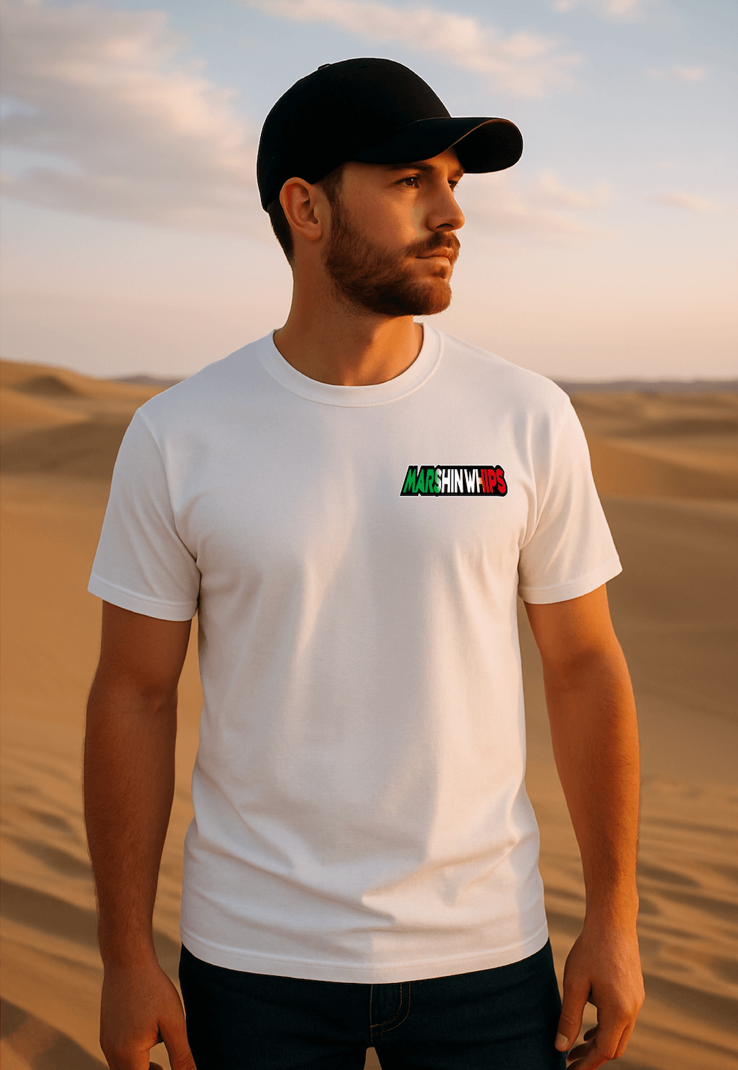Man wearing a white "Marshin Whips" t-shirt with a small front logo in green, white, and red gradient text. He stands in a sandy desert at sunset, looking off to the side with a calm expression under a soft, colorful sky.