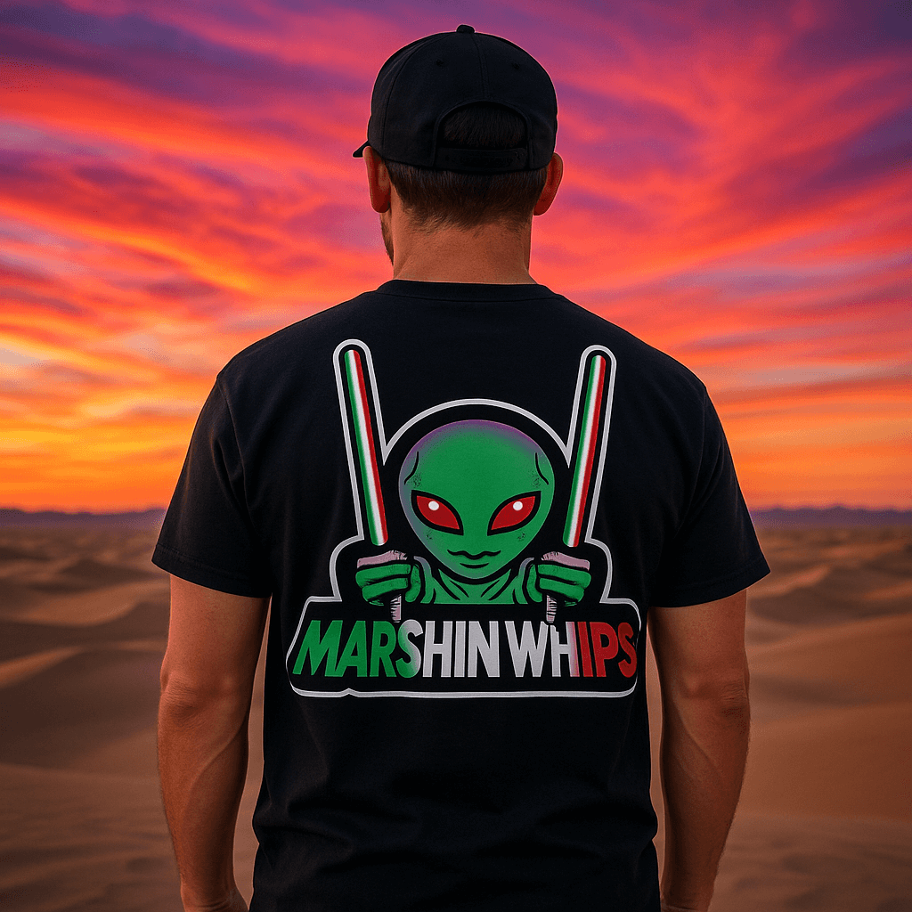 Back view of a man wearing a black "Marshin Whips" t-shirt featuring a vibrant green alien holding red, white, and green light whips for Mexican Pride. He stands in front of a desert landscape with a dramatic sunset sky filled with vivid pink, orange, and purple clouds.