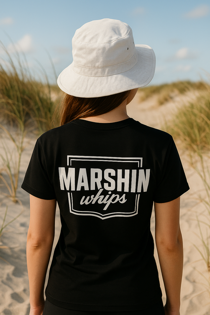 Person wearing a black t-shirt with 'MARSHIN whips' text on a beach